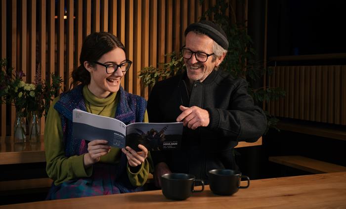 A young woman with dark hair, glasses, a green polo neck and a blue waistcoat sits next to a man with a dark hat, glasses and dark jacket as they look